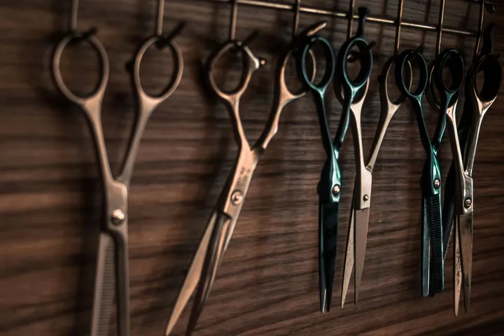 Barber scissors of mixed lengths hanging from a rack on a dark wood wall