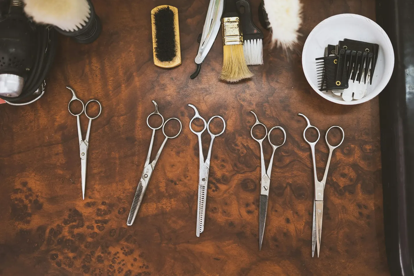 Five barber shears, a neck duster and a razor on a walnut bench.